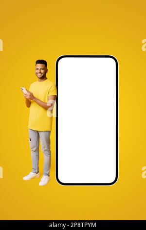 Handsome happy indian guy standing near big huge smartphone with blank screen, copy space, mockup, full length latin man in yellow t-shirt using mobile phone, texting near big phone Stock Photo