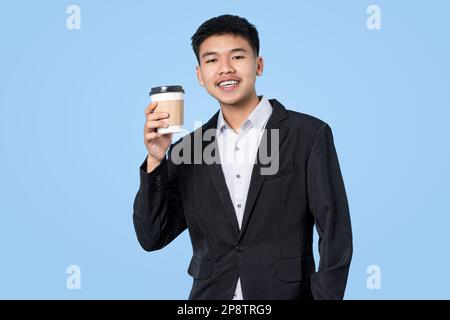 Young handsome asian businessman smiling while holding hot coffee in takeaway paper cup isolated on blue light background. Business Person concept. Stock Photo