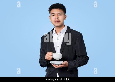 Young handsome asian businessman smiling while holding hot coffee in takeaway paper cup isolated on blue light background. Business Person concept. Stock Photo