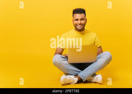 Positive smiling handsome indian man sitting on the floor in lotus pose and using laptop isolated on yellow. Charismatic guy freelancer working remotely, full length, banner Stock Photo