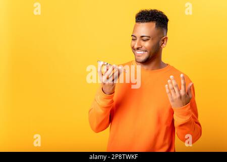 Smiling Indian man is holding smartphone and talking in mic, recording audio message, student guy using assistant, giving commands to artificial intelligence, using voice command Stock Photo
