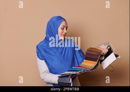 Confident Muslim woman in blue hijab, posing with a catalog of tissues for sofa upholstery on isolated beige background Stock Photo