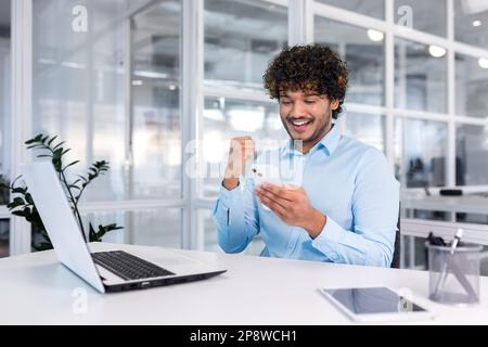Young hispanic businessman sitting in the office at the desk and using the phone. He looks at the screen, dials, rejoices, celebrates, shows a victory gesture with his hand. Stock Photo