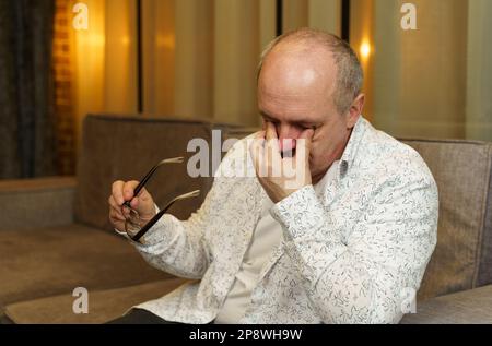 A mature man holds glasses with his hand, the other rubs his eyes. Tired eyes. Stock Photo