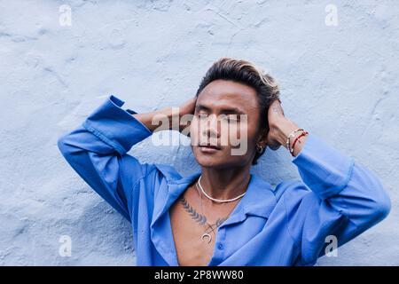 Stylish guy in a blue shirt leaning on a wall touching his head with closed eyes Stock Photo
