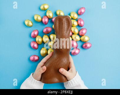 Child's hands holding easter chocolate bunny on blue background with pink and golden candy eggs, easter concept, top view Stock Photo