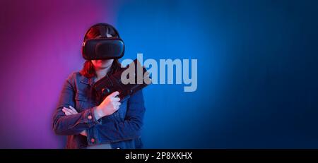 Beautiful young hispanic woman standing front view using virtual reality glasses and game gun in her hands against wall background with blue and purpl Stock Photo