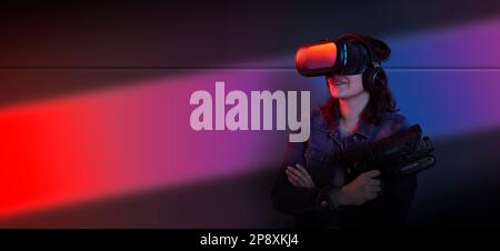 Beautiful young hispanic woman standing using virtual reality glasses and game gun in her hands against wall background with blue and purple light Stock Photo