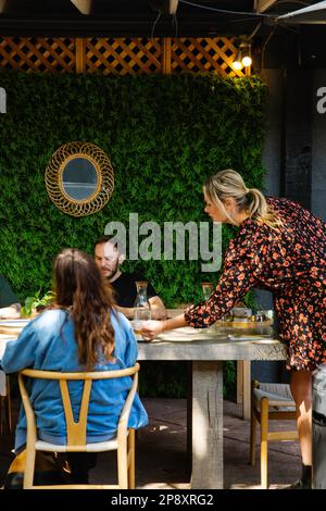 A server delivers dishes to a table in an outdoor dining space with a natural setting Stock Photo