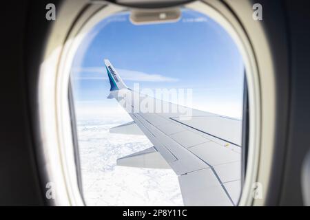 Looking through an airborne WestJet passenger jet window at wing against blue sky and hilly terrain Stock Photo