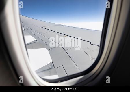 Looking through an airborne passenger jet window at portion of wing, blue sky, and white cloud Stock Photo