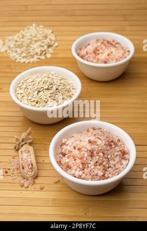 Raw himalayan sea salt and oats are in small white bowls with a small wooden scoop in a studio setting. Stock Photo