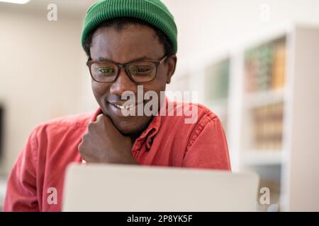 Cheerful happy African guy student sitting in library looking at laptop screen enjoying e-learning Stock Photo