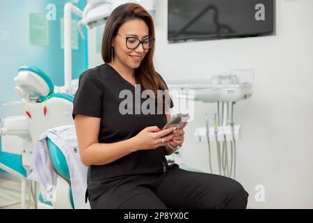 Young Latina dentist sitting in her office, checking her cell phone Stock Photo