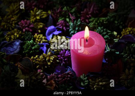 A single, aromatic, vibrant, lit, pink candle surrounded by an assortment of colourful potpourri in soft dark mood lighting and copy space to the left Stock Photo