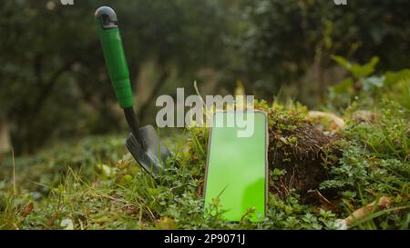 Digital Farming: Small Farmer or Gardener Shovel on Grass with Green Screen Smartphone. Digital Application Concept and Future Agriculture Stock Photo