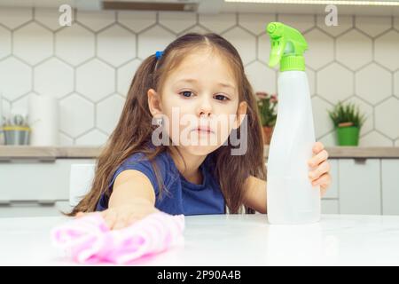 Pretty little girl with detergent sprayer and household rag wipe table. Portrait of child tidying up kitchen, home cleaning concept. Kid help clean up Stock Photo