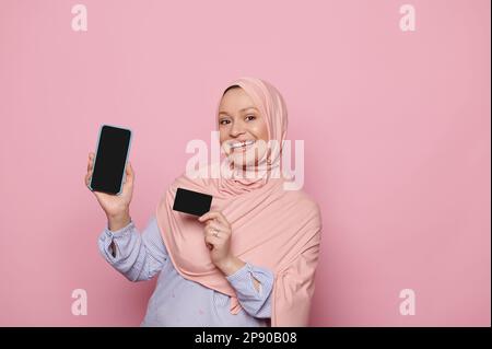 Muslim pretty woman in pink hijab, smiles looking at camera, showing smartphone with blank screen for mobile application Stock Photo