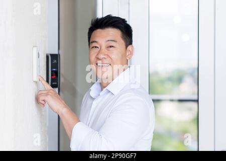 Asian businessman portrait, man walks into modern office, uses doorbell with fingerprint lock, man looks at camera and smiles. Stock Photo