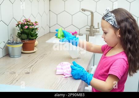 Nice little girl in protective gloves wipe kitchen set with household rag and detergent sprayer. Portrait of child tidying up kitchen side view. Home Stock Photo