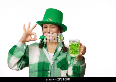 Cheerful woman in green party hat, holds a mug of Irish beer, winks, shows Ok hand sign at camera. Saint Patrick's Day Stock Photo