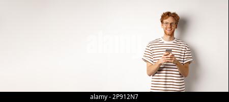 Technology and e-commerce concept. Redhead guy in glasses using mobile phone and smiling. Young man with smartphone staring happy at camera, white Stock Photo