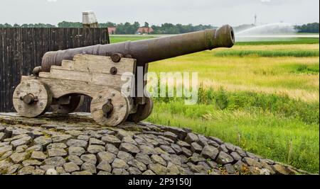 A vintage cannon located on a large rock near the shoreline, with a metal fence to the side Stock Photo