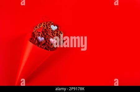 Mini pretzels and small colorful hearts in red paper cone lie on red background. Pretzel sunday concept. Banner with copy space Stock Photo