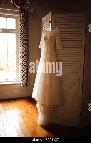 A beautiful white wedding dress is elegantly draped over a wall in a classic and traditional house, creating a timeless and romantic atmosphere Stock Photo