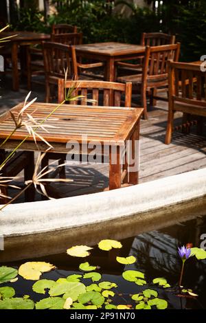 Outdoor cafe patio with old, shabby wooden tables and chairs in the sunlight. Pond with water lilies on a  romantic restaurant terrace Stock Photo