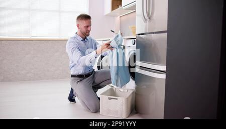 Young Man Loading Clothes Into Washing Machine In Kitchen Stock Photo
