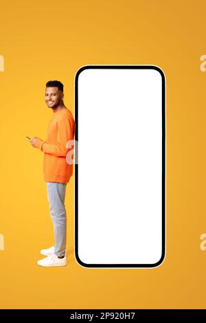 Smiling attractive guy standing near huge smartphone with empty blank screen, man with phone in his hands advertising new app or website, mockup Stock Photo