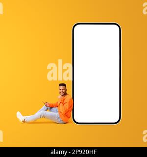 Excited Indian young man sitting on the floor near huge smartphone and points finger at empty screen isolated on yellow background, arab guy presenting and advertising mobile app, copy space Stock Photo