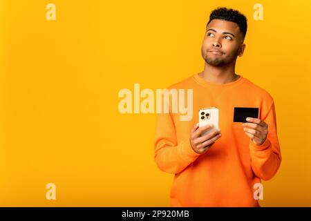 Middle-eastern positive man is thinking about earning online. Thoughtful handsome indian guy holding credit card and smartphone, dreaming about long-awaited purchase standing isolated on yellow Stock Photo
