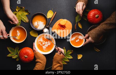 Flat-lay of family or friends enjoying dinner with pumpkin cream soup in plates on black background decorated with pumpkins and fall yellow leaves. Stock Photo
