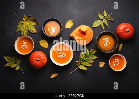 Flat-lay of dinner with homemade warming pumpkin cream soup in plates on black background decorated with pumpkins and fall yellow leaves. Autumn Stock Photo