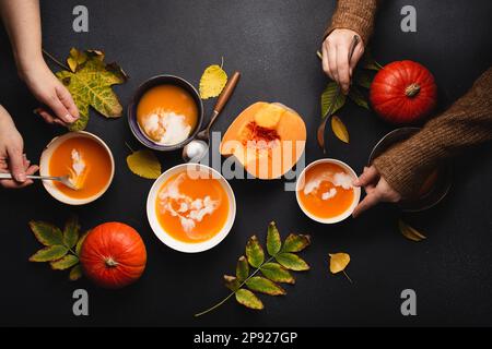 Flat-lay of family or friends enjoying dinner with pumpkin cream soup in plates on black background decorated with pumpkins and fall yellow leaves. Stock Photo
