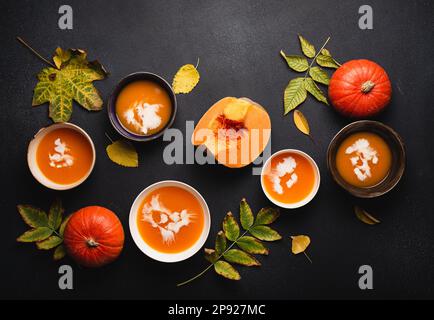 Flat-lay of dinner with homemade warming pumpkin cream soup in plates on black background decorated with pumpkins and fall yellow leaves. Autumn Stock Photo