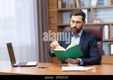 A young man is sitting in the office in a suit at the table, holding and concentrating on reading a green book. Resting, studying, break. Stock Photo