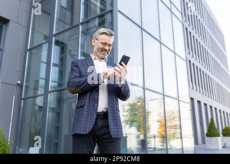 Happy senior gray-haired businessman, lawyer, investor stands outside the office, skyscraper. Joyful uses the phone, reads, sends, received. Stock Photo