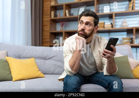An upset young man is sitting on the sofa at home with a phone in his hands, looking thoughtfully to the side. Waiting and received a message from a girlfriend, lover, ex, wife, Stock Photo