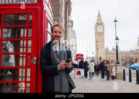 Smiling man with a smartphone in his hands stands near the famous red telephone booth on the street of London Stock Photo