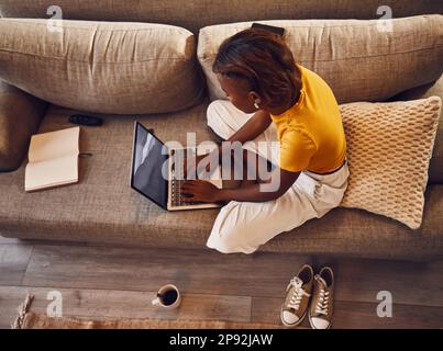Distance learning student writing, studying and typing on a laptop doing a virtual assignment test on the sofa at home. Above view of one young female Stock Photo