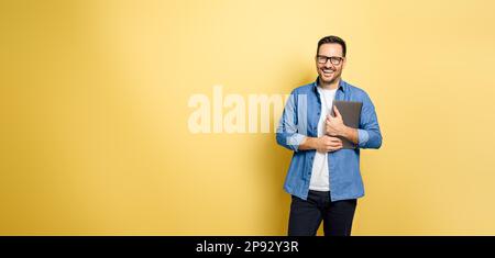 Portrait of cheerful young adult man holding laptop and standing against yellow background. Happy male freelancer looks at camera wearing eyeglasses. Stock Photo
