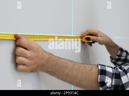 Man using cross line laser level and tape for accurate measurement on light wall, closeup Stock Photo