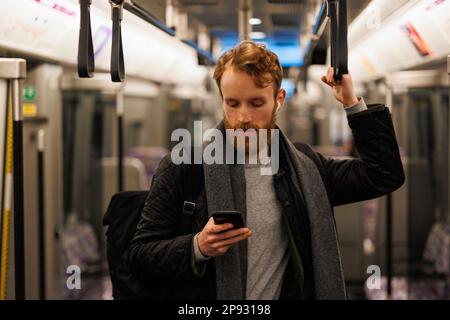 Stylishly dressed bearded man stands in a subway car in headphones and a smartphone in his hand. Passenger in public transport Stock Photo