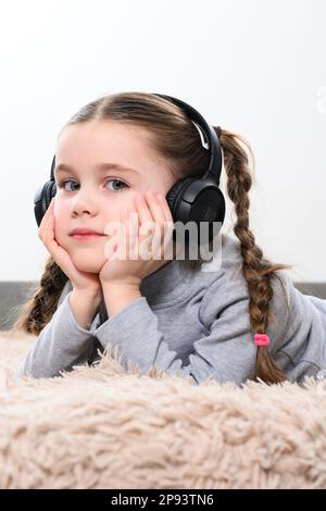 Ivano-Frankivsk, Ukraine January 28, 2023: little girl listens to songs through wireless headphones and lies on the sofa, wireless headset accessory Stock Photo