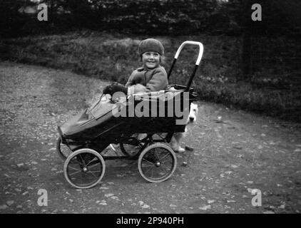 circa 1930s, historical, outside on a bumpy path, a sweet young girl, with a big smile, sitting in a steel-framed folding pushchair holding a lead for her scottie dog which is hiding behind the child's transport, England, UK. Stock Photo