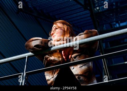 Young woman with reddish hair and glasses in plaid shirt jacket leans over roofed railing and looks into distance, seen from below Stock Photo