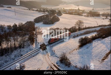 Germany, Thuringia, Allendorf (background), regional train 60, train 29887, level crossing, country lane, landscape, village, fields, forest, snow, overview, oblique view, evening light, back light Stock Photo